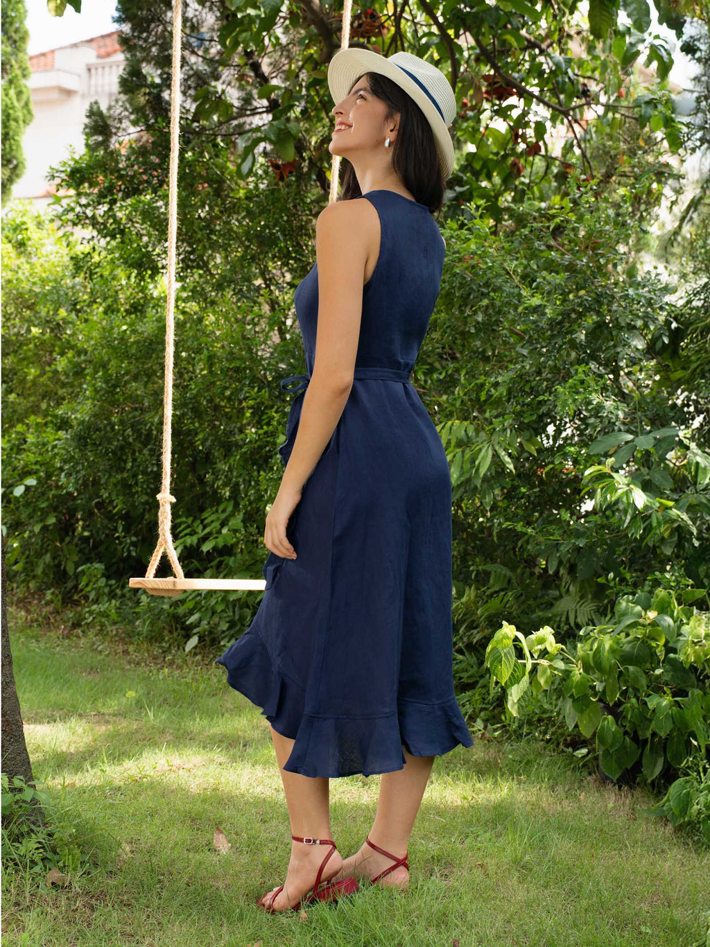 Woman in a blue dress and white hat standing in a garden with a swing in the background.