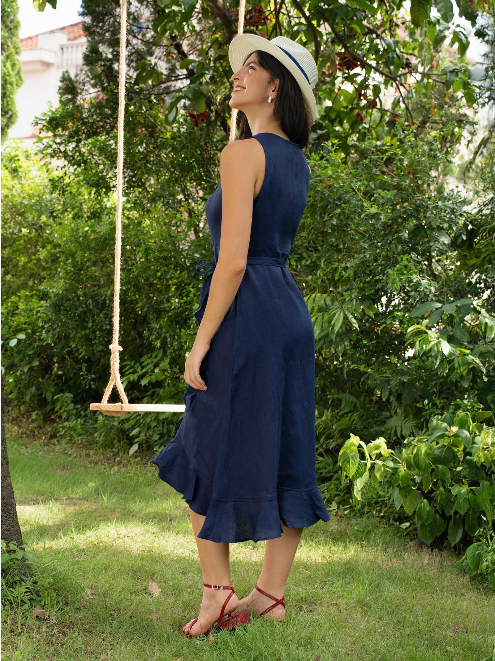 Woman in a blue dress and white hat standing in a garden with a swing in the background.