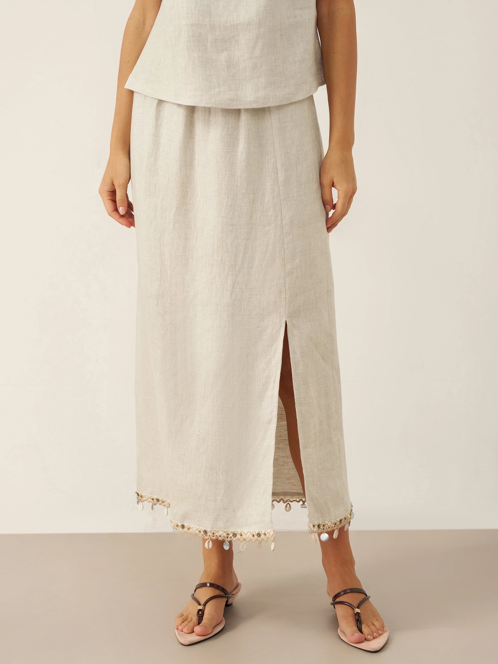 Person wearing a light-colored linen skirt with a slit and decorative elements on a plain background