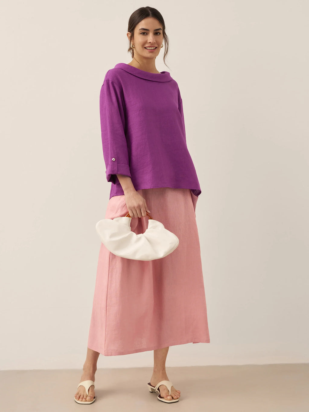 Woman wearing a purple top and pink skirt holding a white clutch against a plain background