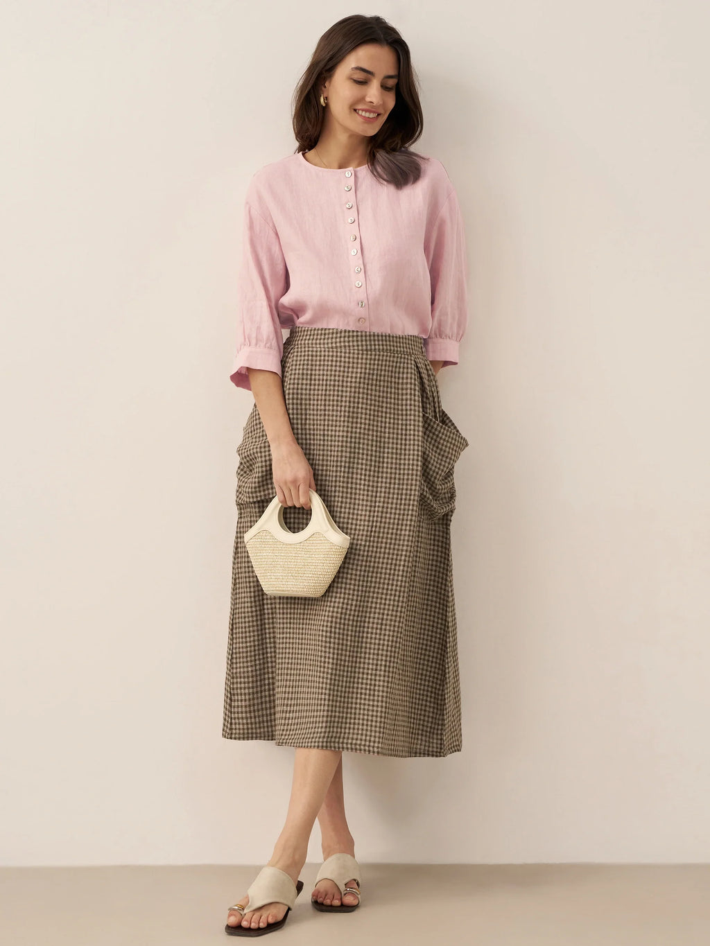 Woman wearing a pink blouse and checkered skirt holding a white handbag against a plain background