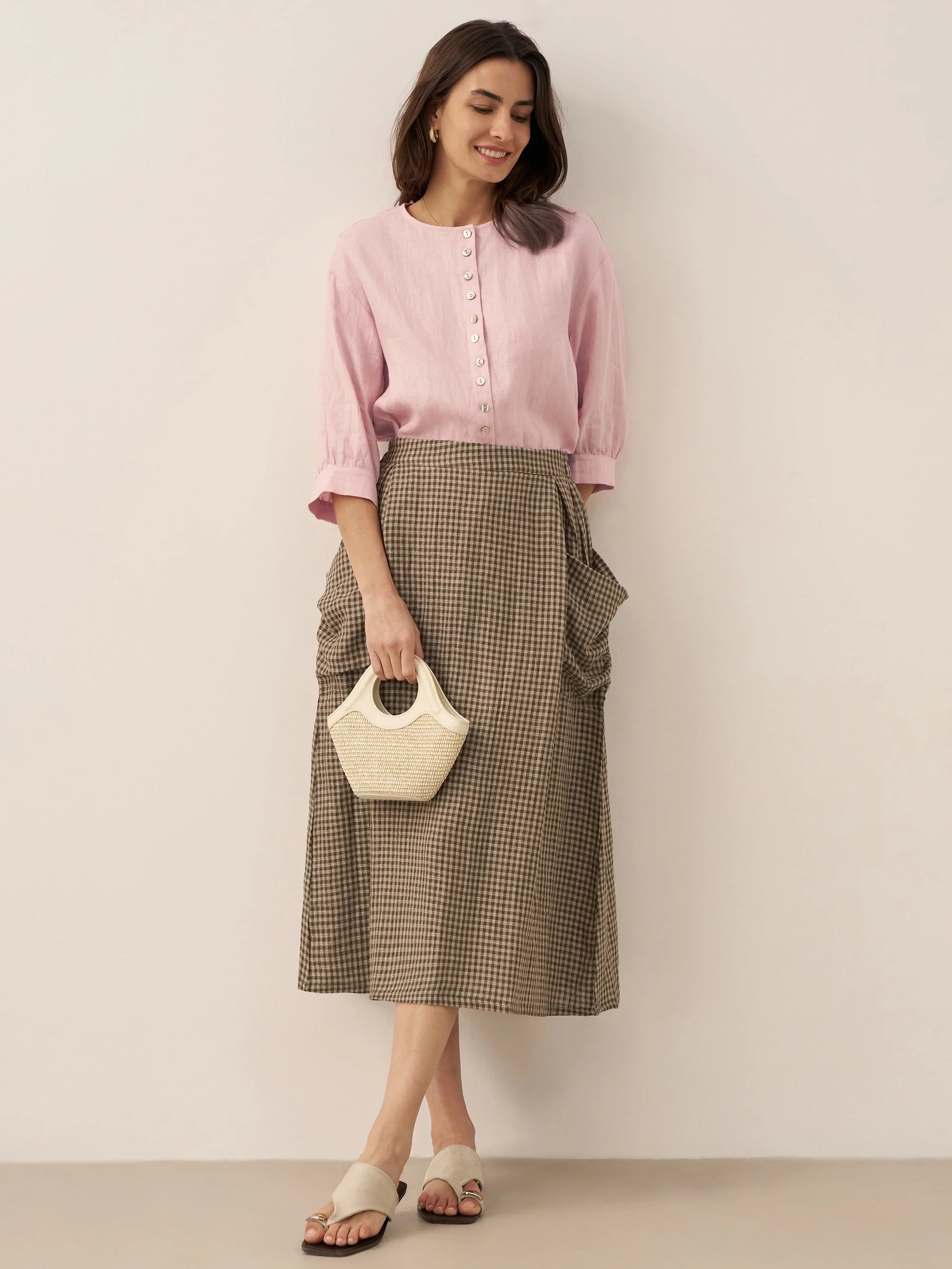 Woman wearing a pink blouse and checkered skirt holding a white handbag against a plain background