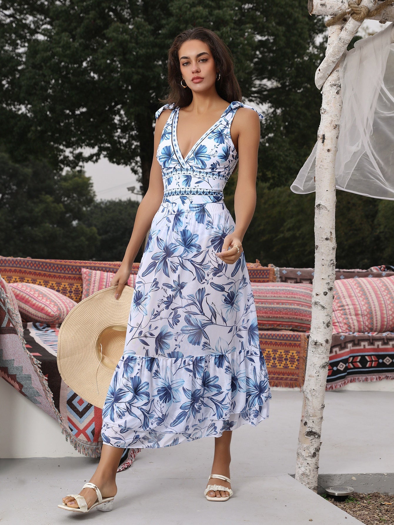 Woman in a blue floral dress holding a sun hat, standing in front of outdoor furniture.