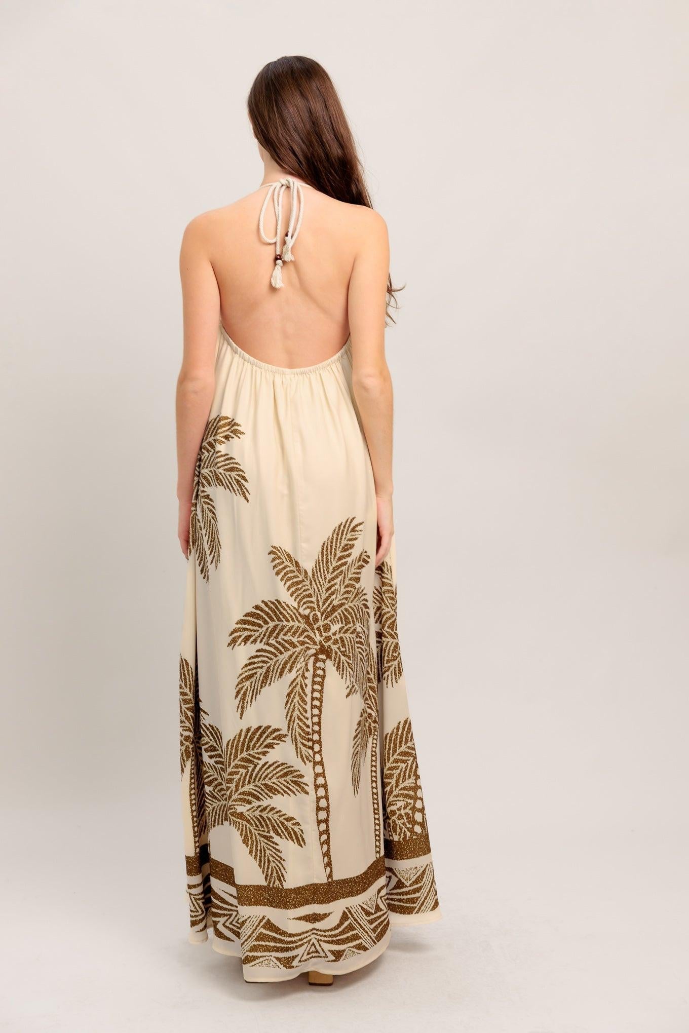 Woman wearing a beige maxi dress with palm tree patterns on a plain background