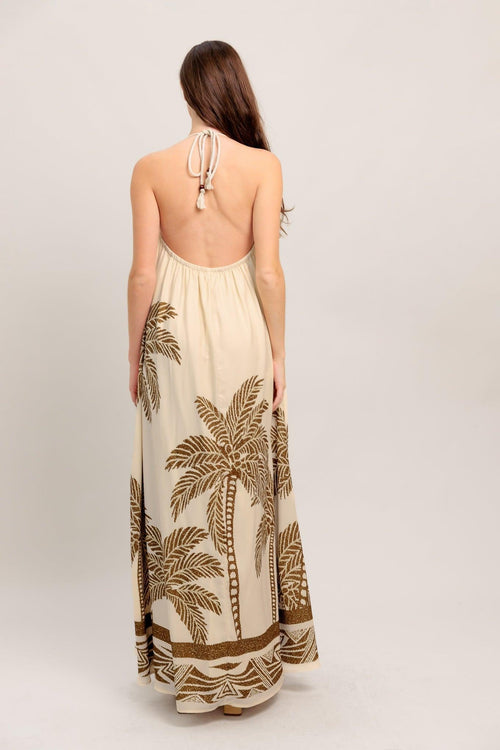 Woman wearing a beige maxi dress with palm tree patterns on a plain background