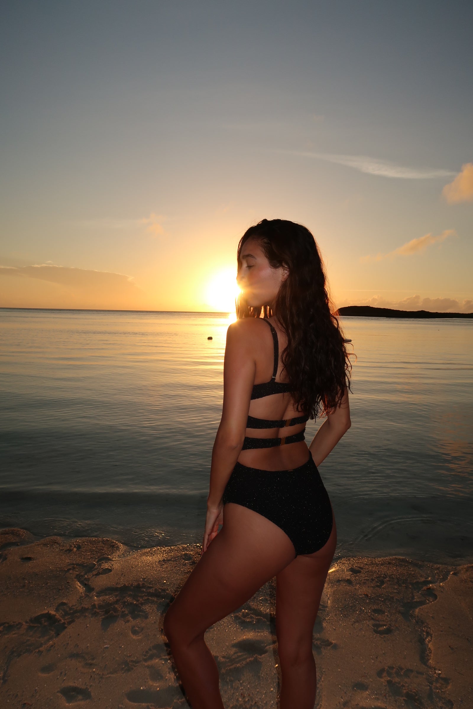 Woman in a black swimsuit standing on a beach at sunset.