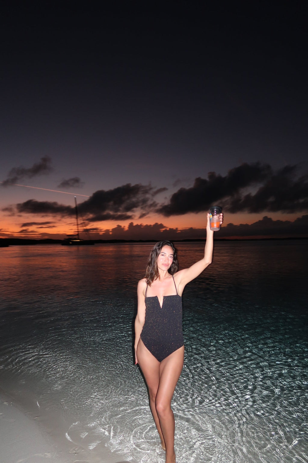 Woman in a black swimsuit holding a drink on a beach at sunset.