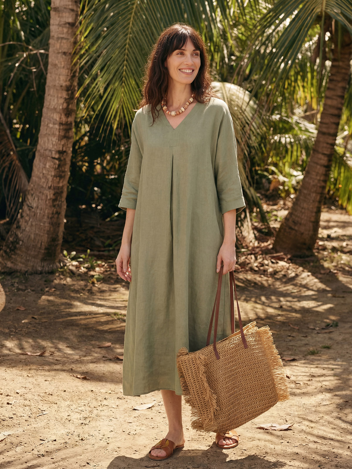 Woman in a green linen dress holding a woven bag in a tropical setting with palm trees.
