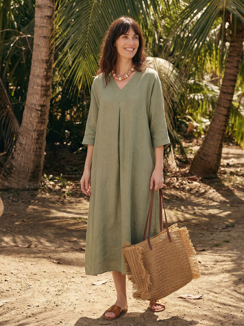 Woman in a green linen dress holding a woven bag in a tropical setting with palm trees.